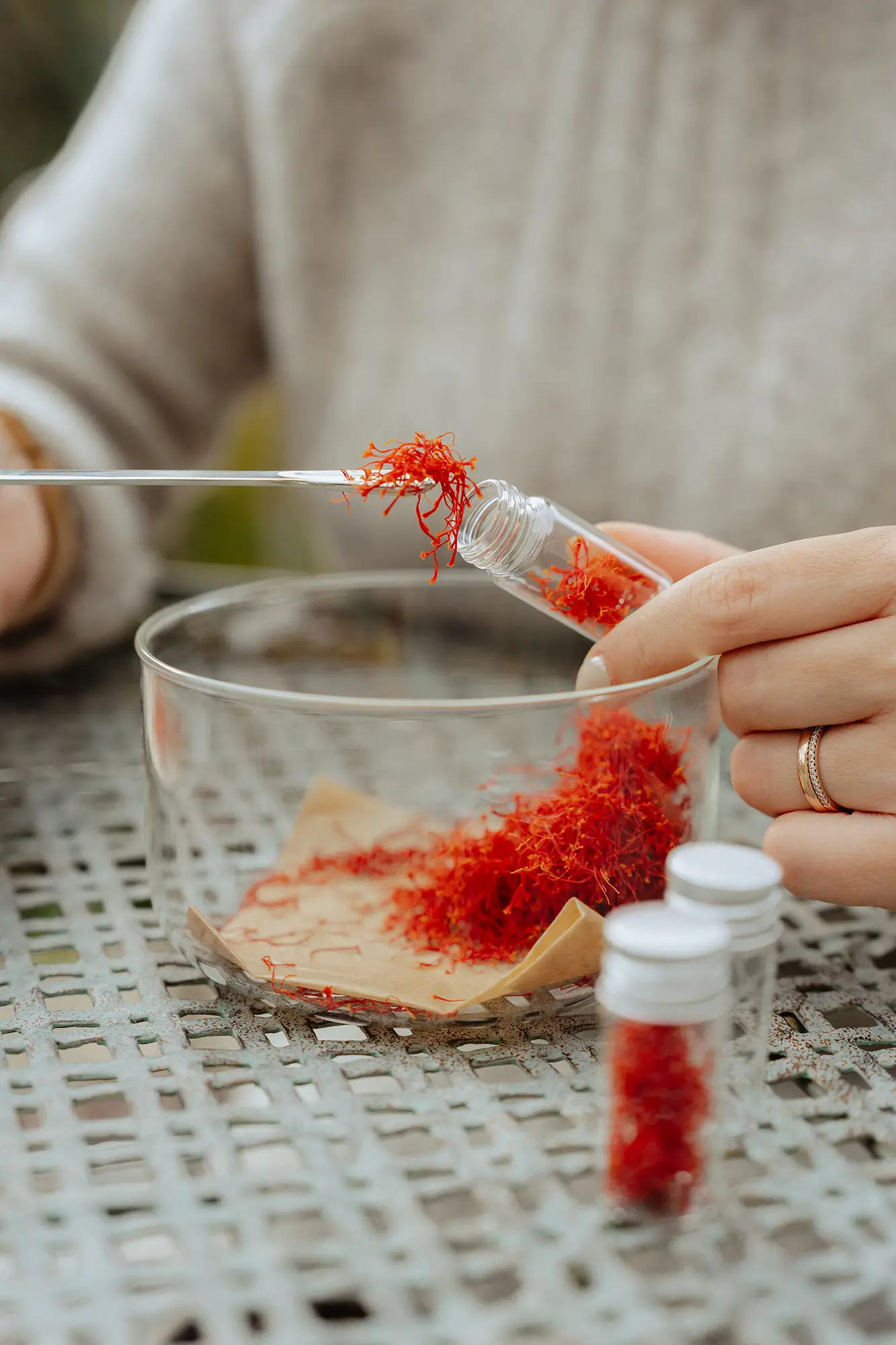 Safranfäden werden mit einer Pipette sorgfältig in ein kleines Glasröhrchen abgefüllt; im Vordergrund liegen weitere Safranfäden auf Papier in einer Schale, während eine Hand das Gefäß hält.