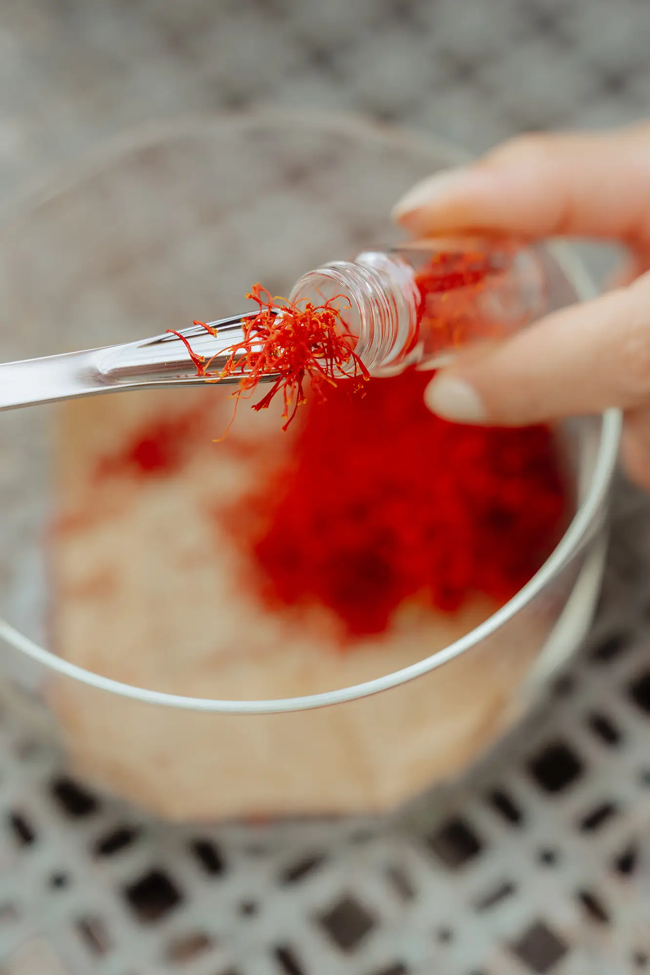 Safranfäden werden mit einer Pipette vorsichtig in ein kleines Glasgefäß gefüllt; im Hintergrund liegen weitere rote Safranfäden unscharf in einer Schale.
